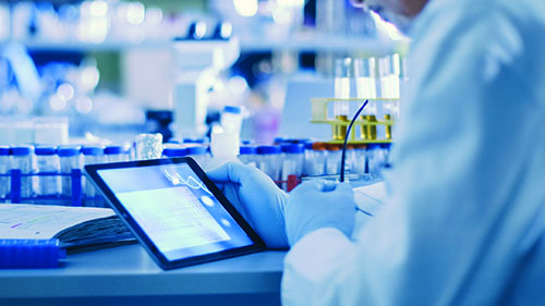 A laboratorian sitting at a table with lab equipment and looking at a tablet.