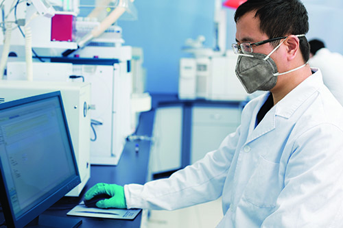 A male laboratorian in a mask and lab coat sitting and working on a computer