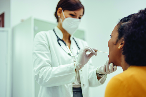 A woman doctor in a mask hold a swab inside a woman patient's mouth.