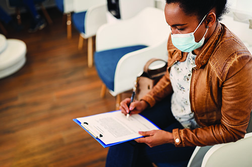 A black woman in a mask sitting in a waiting room filling out a form on a clipboard.