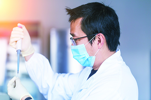 A male laboratorian wearing glasses, a blue mask, and a lab coat, insterting something from a pipette to a test tube.