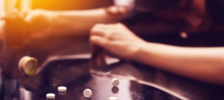 Pills lay scattered on a counter; a person’s arms partially blurred in the background also rests on the counter