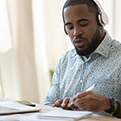 An African American man wearing headphones and facing a computer writes in a notebook on his left side.