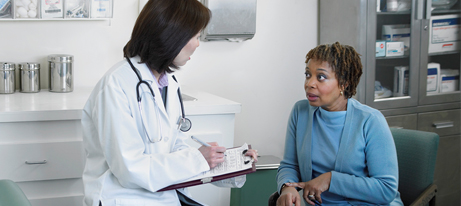 A female doctor writing notes on papers on a clipboard she is holding speaks to a female patient, who is sitting in a chair.