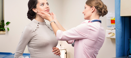 A standing female physician holds her hands on the neck of a pregnant female patient, who also is standing